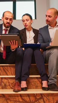 Team of business workers making plans of work, using the laptop and tablet, enjoying sitting on staircase explaining finance project. Group of company manager and office executive on busy stairs