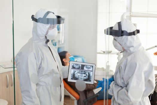 Dentist doctors with ppe suit analysing teeth x-ray using tablet in dental room, planning surgery during global pandemic while patient waiting on stomatological chair.