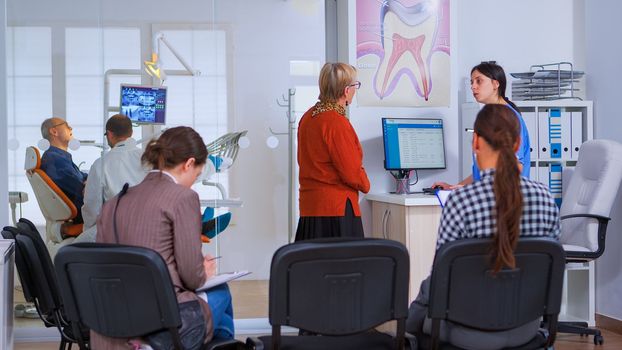 Young lady visiting stomatological clinic for teeth checking while dentistry doctor preparing old man for dental surgery in background. Patients sitting in crowded waiting room of orthodontist office