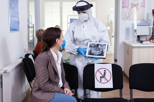 Stomatologist in protective suit pointing on digital x-ray of tooth explaining to patient treatment using tablet in covid-19 pandemic. Medical team wearing face shield, coverall, mask and gloves.