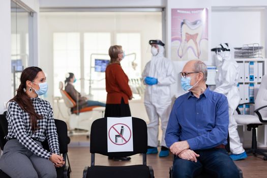 Senior man with face mask discussing with woman patient in stomatology clinic in waiting room, keeping social distancing during global pandemic with coronavirus. Doctor standing wearing ppe suit.