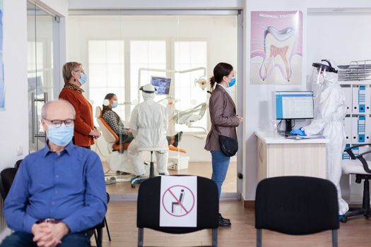 Patients with protection mask waiting in stomatology reception with new normal, staff wearing ppe suit. Woman checking appoiment with nurse coming for dental control during coronavirus pandemic