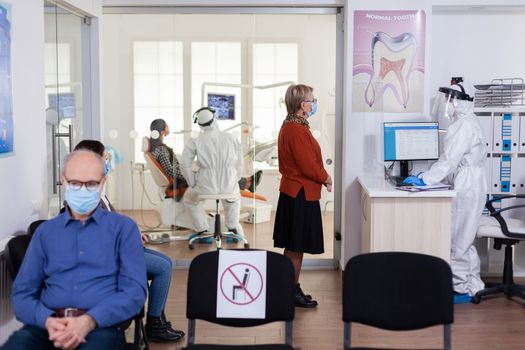 Dentist doctor in ppe suit consulting patient in dental clinic dressed in ppe suit as safety precation during global pandemic with coronaivurs. Senior woman talking with dentistiry stomatology receptionist sitting on chair.