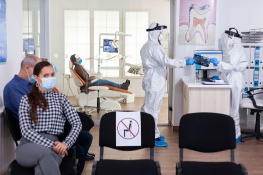 Stomatology dentist dressed in ppe suit taking patient x-ray from receptionist in clinic hallway, keeping social distancing during global pandemic with coronavirus.