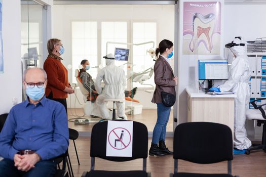 Patients with protection mask waiting in stomatology reception with new normal, staff wearing ppe suit. Woman checking appoiment with nurse coming for dental control during coronavirus pandemic