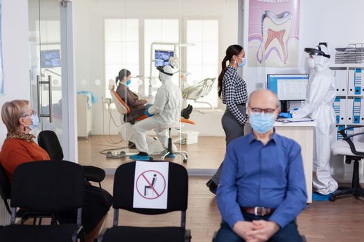 Dentist nurse dressed in ppe suit with face shiled discussing with patient in stomatology waiting room. People keeping social distancing as prevention during coronavirus outbreak.