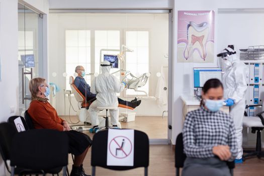 Doctor in pee suit consulting senior patient in stomatology office keeping visor, during global pandemic with coronavirus, new normal. Receptionist with face mask in dentistiry waiting area.