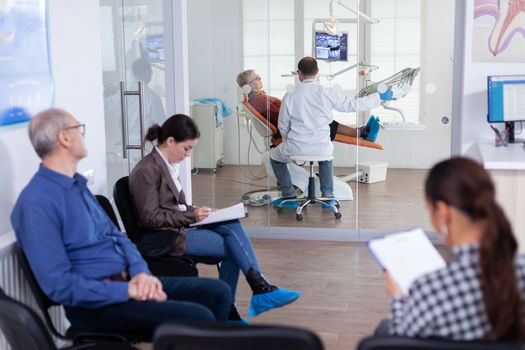 Crowded stomatology waiting area with people filling form for dental consultation. Stomatoloy specialist denstiry treating senior woman cavity. Receptionist working on computer.