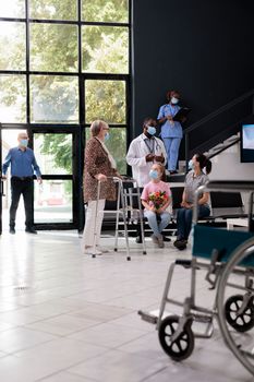 Senior woman with walking frame discussing disease diagnosis with doctor during checkup visit in hospital reception. People wearing medical protective face mask to prevent infection with coronavirus