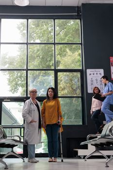 Senior doctor helping asian patient with leg injury to use walk frame, dealing with physical impairment after fracture injury. Injured woman doing physiotherapy rehabilitation with walking aid.