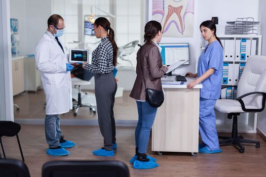 Dentist doctor teeth health care specialist explaining treament to patient in stomatology waiting area pointing at tablet pc with radiography. People filling form in dentistiry reception.