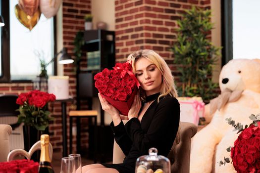 Attractive woman holding valentines day red roses bouquet, receiving presents from boyfriend during love holdiday. Cute girl sitting in living room filled with macarons cookies and romantic presents