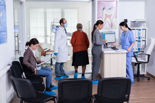 Young lady asking informations filling in stomatological form while patients talking sitting on chair in waiting area. People speaking in crowded professional orthodontist reception office.