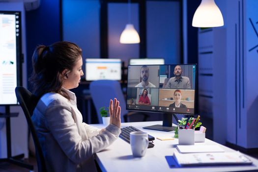 Back view of business woman talking about sale report in video conference working overtime in start-up office. Team using online wireless communication discussing on webcam during group meeting.