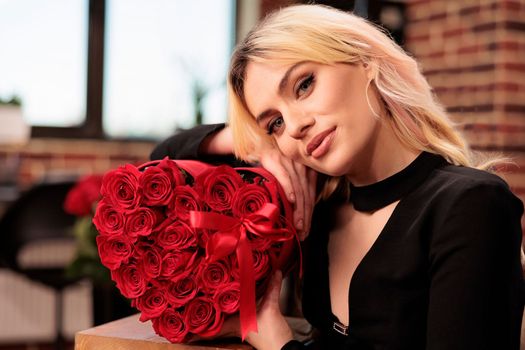 Girlfriend standing in living room filled with romantic presents, holding red roses in heart shaped box. Beautiful woman in elegant black dress enjoying celebrating valentine s day. Love concept