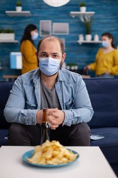 Man with protection mask looking at camera holding bottle of beer ready for party with friends in living room respecting social distance in global pandemic. People socializing during covid 19 outbreak