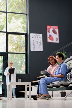 Medical asisstant consulting senior patient in hospital waiting area, filling report papers explaining healthcare treatment. Nurse doing checkup examination with asian woman in lobby.