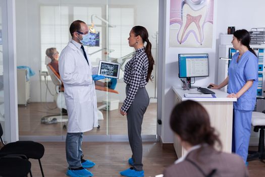 Dentist with face mask showing patient dental x-ray in stomatology specialist reception. Explaining diagnosis during appointment, senior man sitting in dental chair.