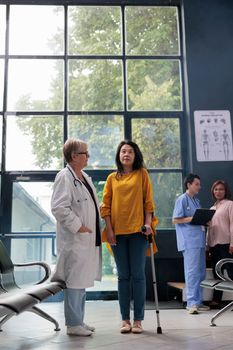 Injured patient trying to walk with walking cane in hospital lobby, receiving help from senior physician. Old woman doing physiotherapy exam for physical injury rehabilitation. Medicine support