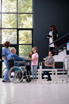 Little girl giving bouquet of flowers to disabled grandfather during checkup visit appointment in hospital waiting room. Patient in wheelchair discussing health care treatment with medical assistant