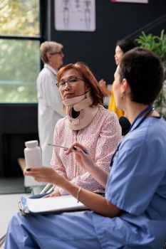 Senior patient with cervical neck collar having medical appointment in hospital waiting area. Specialist holding pills bottles explaining medication prescription. Medicine support