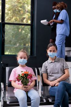 Portrait of family sitting on chair in hospital waiting lobby during checkup visit appointment, wearing medical face mask to prevent infection with covid19. Little girl holding flowers bouquet.
