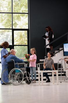 Granddaughter giving bouquet of flowers to disabled grandfather during checkup visit appointment in hospital waiting area. Patient in wheelchair discussing medication treatment with medical nurse