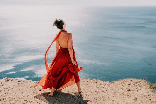 Side view a Young beautiful sensual woman in a red long dress posing on a rock high above the sea during sunrise. Girl on the nature on blue sky background. Fashion photo.