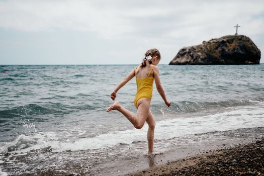 Cute little girl running along the seashore against a clear blue sea and rejoices in the rays of the summer sun. Beautiful girl in yellow swimsuit running and having fun on tropical beach