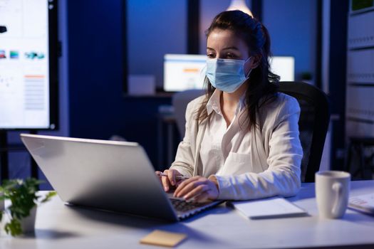 Close up of businesswoman with face mask checking emails late at night in new normal business office before deadline. Taking notes, analysing documents overtime during global pandemic