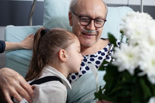 Girl hugging old man, lying in hospital bed. Senior, granddaughter showing physical affection each other in geriatric clinic observation room. Happy elderly patient receiving surprise from child.