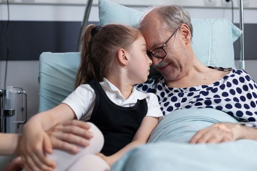 Elderly recovering patient happily cuddling infant granddaughter in hospital observation room bed. Senior man enjoying company of relatives visit in geriatric clinic room.