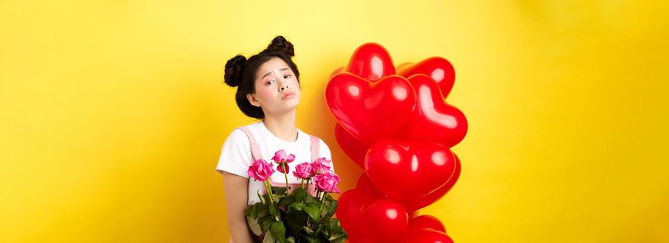 Happy Valentines concept. Sad and gloomy asian woman holding bouquet of roses and feeling upset and lonely on romantic lovers day, standing near red heart balloons, yellow background.