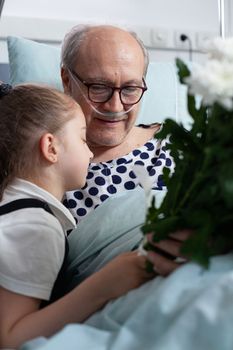 Little girl giving flowers bouquet present, hugging grandfather admitted to hospital. Elderly bedridden patient saying goodbye small child in geriatric clinic visiting room.