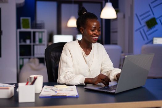 Smiling african american businesswoman planning management annual graph typing company strategy on laptop computer late at night in startup office. Black manager browsing corporate information