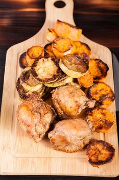 Top view of wooden board with homemade fried chicken next to grilled vegetables on wooden background