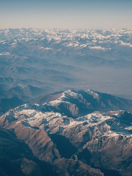 Smooth outline of the Alps from above. Alpine mountain system stretches to the horizon line, the slopes are covered with snow caps in some places, the sun's rays illuminate the slopes
