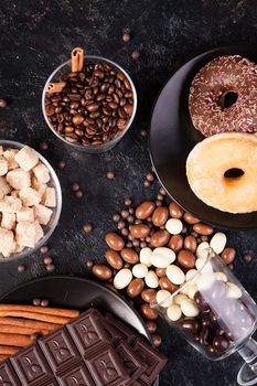 Top view of peanuts in chocolate, spilled on the dark board, next to chocolate tablets, donuts, brown sugar and coffee beans. Studio photo