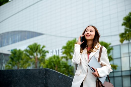 Happy successful Asian businesswoman holding laptop and Talk with cellphone on the Street next to a glass building. successfully work go to against city scene in background of modern office buildings.