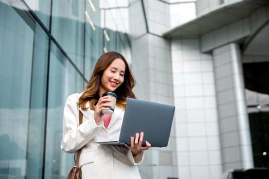 Confident businesswoman using computer laptop standing while drinking a cup of coffee takeaway at the city exterior. Beautiful smile female being ready to work. Business lifestyle concept.