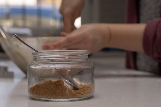 glass jar with brown sugar and a spoon in the background the hands of a woman preparing a cake