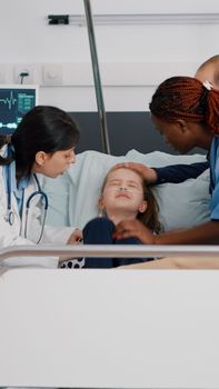 Pediatrician woman doctor holding sick little child monitoring disease symptom during sickness examination in hospital ward. Hospitalized kid recovering after suffering medical surgery