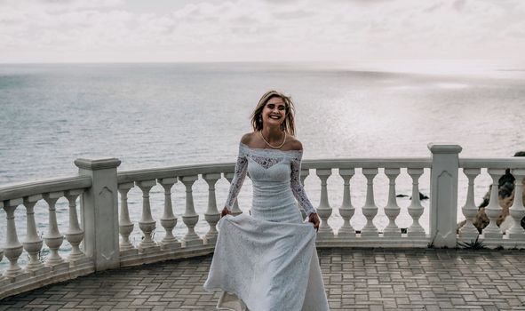 Romantic beautiful bride in white dress posing with sea and mountains in background. Stylish bride standing back on beautiful landscape of sea and mountains on sunset