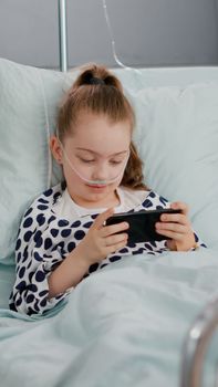 Sick little child resting in bed playing online video games using smartphone relaxing after suffering sickness recovery surgery. Kid wearing nasal tube during medical examination in hospital ward