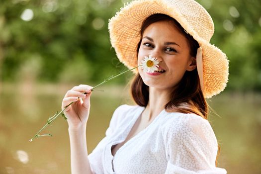 close-up portrait of a happy woman in a wicker hat with a camomile in her hands on the background of the lake. High quality photo