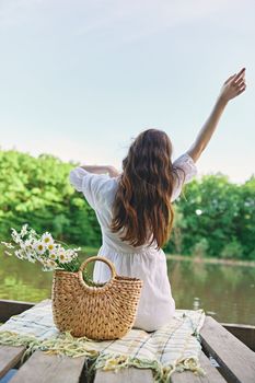 a woman sits on a blanket near the lake with her hands raised above her head and looks at the view. High quality photo