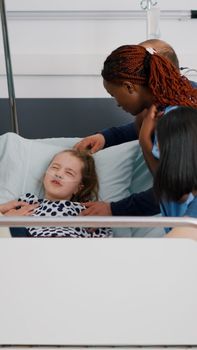 Hospitalized sick child suffering pain attack while medical team holding her during sickness examination in hospital ward. Pediatrician african american nurse trying to listening oxygen saturation