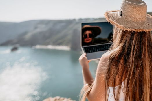 Successful business woman in yellow hat working on laptop by the sea. Pretty lady typing on computer at summer day outdoors. Freelance, travel and holidays concept.