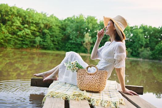 happy woman in a long light dress is resting by the lake sitting on a plaid and enjoying the view. High quality photo