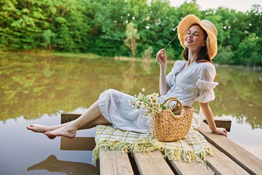 a woman sitting on a pier by the lake in a light dress and a wicker hat smells a chamomile flower. High quality photo